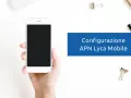 Configurazione APN Lycamobile Italia: mano che tiene uno smartphone bianco con schermo nero, su sfondo bianco con graffette.