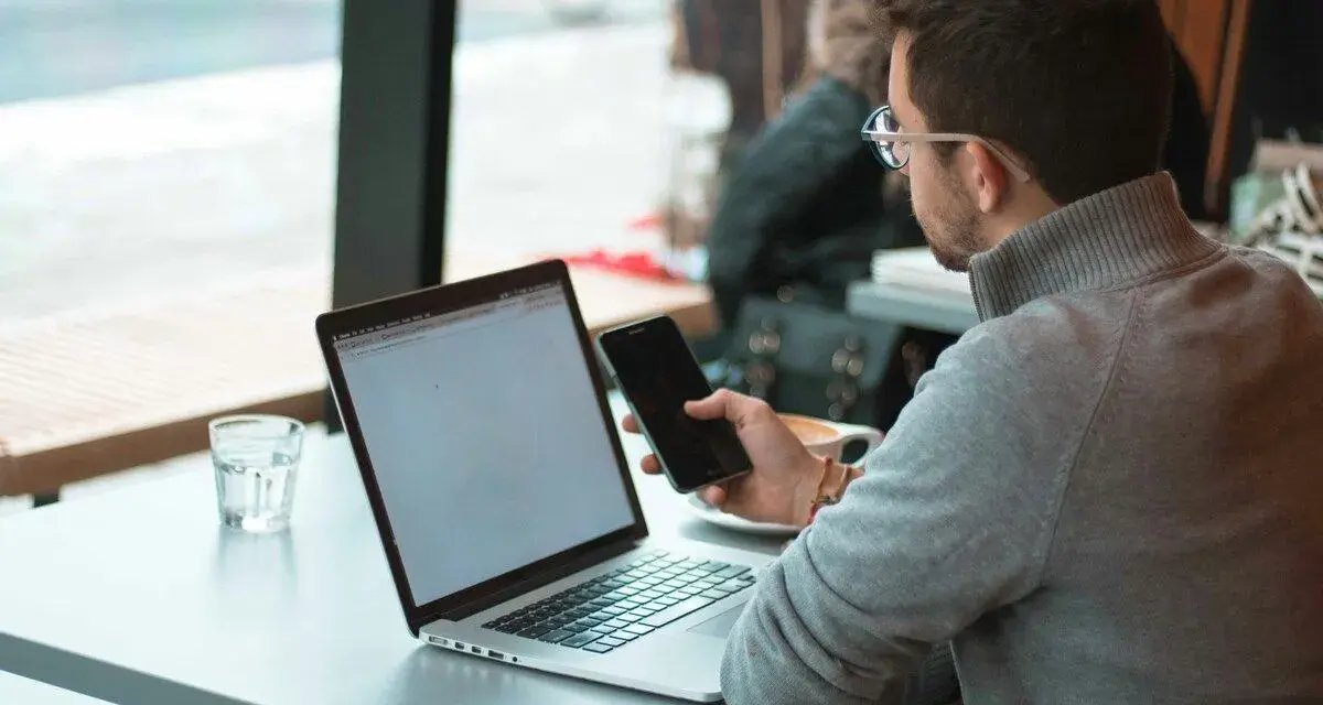 Uomo con occhiali che usa il telefono e il laptop, scopri come collegare il telefono al pc senza cavo per una maggiore comodità.