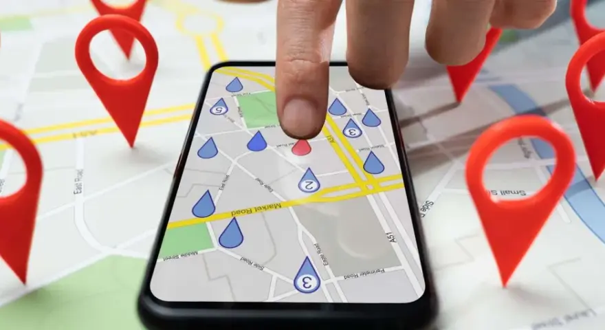 Dita la mappa con puntatori rossi, un dito tocca uno smartphone che mostra una mappa con gocce d'acqua, simulando gli spostamenti su Google Maps.