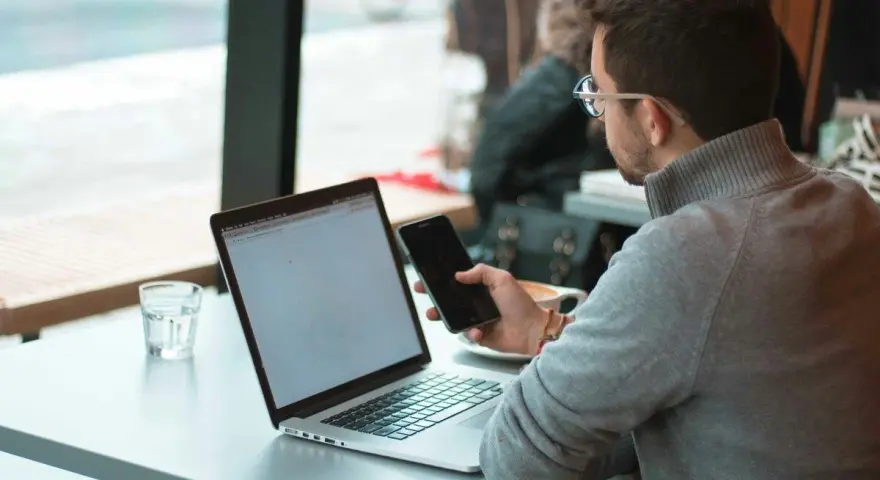 Uomo con occhiali che usa il telefono e il laptop, scopri come collegare il telefono al pc senza cavo per una maggiore comodità.