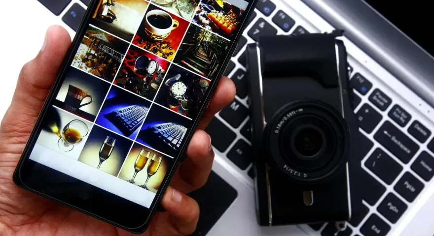 Smartphone con galleria foto su tastiera laptop, accanto a fotocamera. Guida rapida per scaricare le foto dal cellulare al pc.